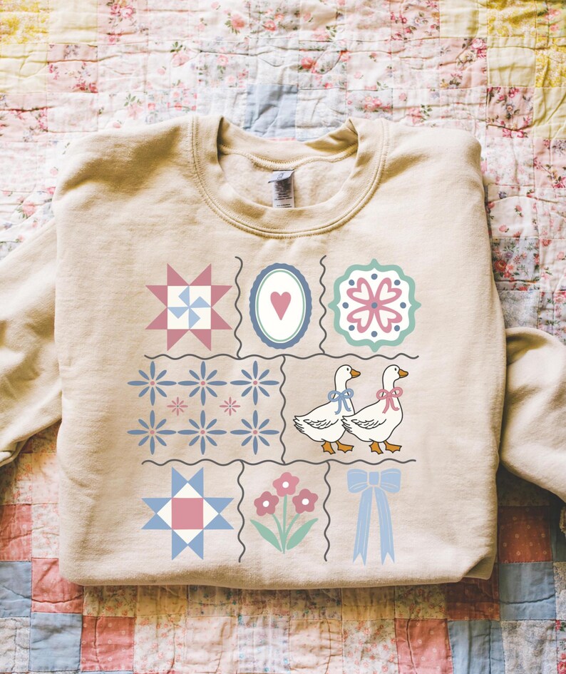 May include: A beige sweatshirt featuring a patchwork design with various quilt block patterns, including stars, flowers, and hearts. Two geese with blue and pink bows are also featured. The sweatshirt has a crew neck.