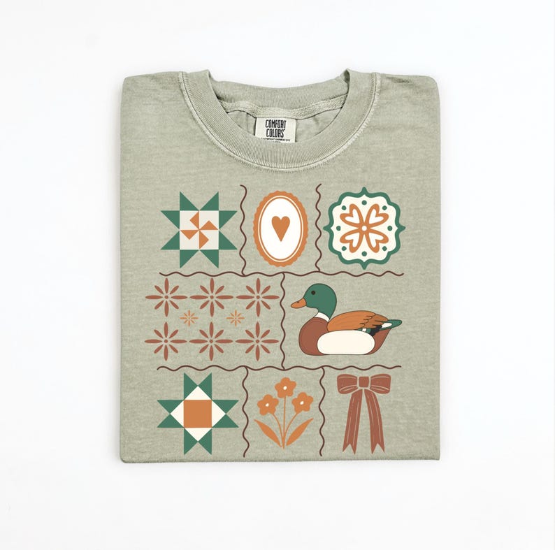 May include: Sage green t-shirt with a grid design featuring a duck, flowers, stars, and a bow. The shirt has a crew neck and short sleeves. The Comfort Colors brand is visible on the collar.