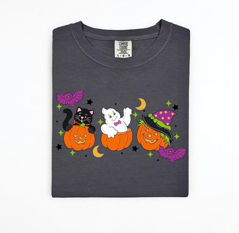 May include: Dark gray t-shirt featuring a Halloween design. The design includes a black cat, a ghost, and a jack-o'-lantern, each in a carved pumpkin. Purple bats, stars, and crescent moons complete the festive look.