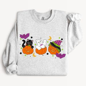 May include: A light gray sweatshirt featuring Halloween-themed graphics. The design includes a black cat, a ghost, and a pumpkin wearing a witch hat, each in a carved pumpkin. Purple bats, stars, and moons complete the festive look.