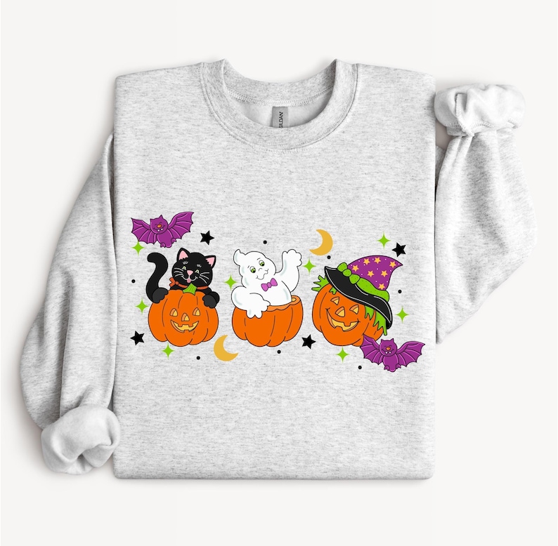 May include: A light gray sweatshirt featuring Halloween-themed graphics. The design includes a black cat, a ghost, and a pumpkin wearing a witch hat, each in a carved pumpkin. Purple bats, stars, and moons complete the festive look.