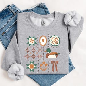 May include: A light gray sweatshirt with a colorful patchwork design featuring a duck, hearts, flowers, and geometric shapes. The sweatshirt is laid on top of blue jeans.