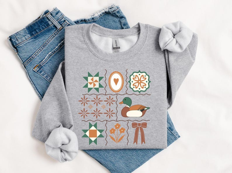 May include: A light gray sweatshirt with a colorful patchwork design featuring a duck, hearts, flowers, and geometric shapes. The sweatshirt is laid on top of blue jeans.
