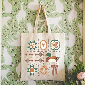 May include: A cream-colored tote bag with a patchwork design featuring geometric shapes, a duck illustration, and a bow. The bag is hanging against a green floral wallpaper with white cat silhouettes.