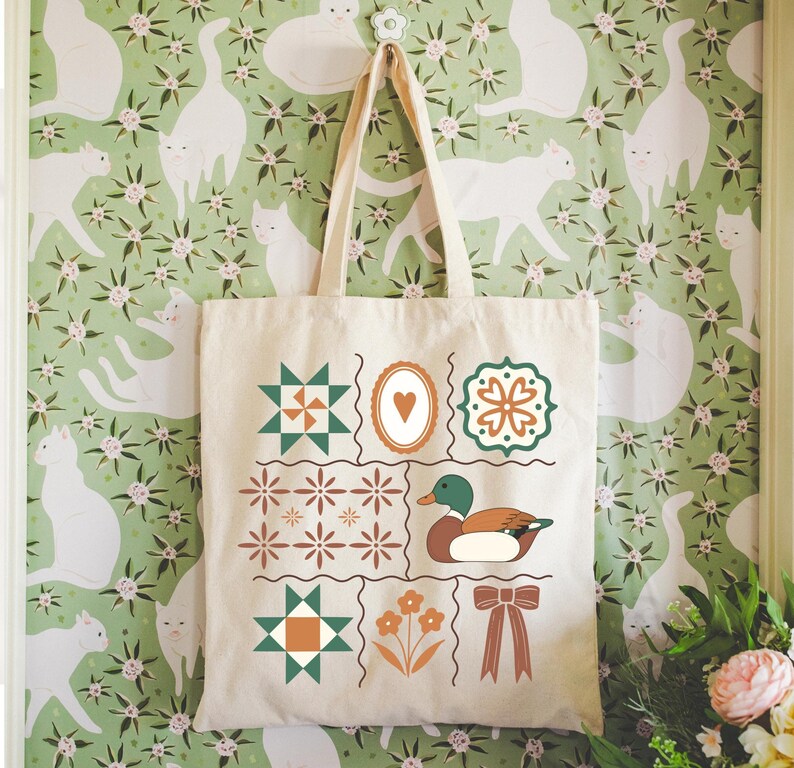May include: A cream-colored tote bag with a patchwork design featuring geometric shapes, a duck illustration, and a bow. The bag is hanging against a green floral wallpaper with white cat silhouettes.
