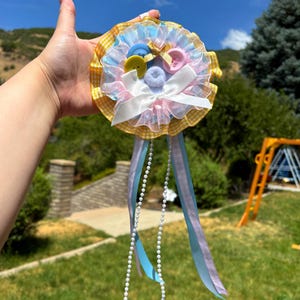May include: A circular rosette badge featuring a yellow and white gingham outer border, layered with light blue and pink ruffled organza. The center is decorated with blue, yellow, and pink buttons, a small gold ribbon, and a white satin bow.