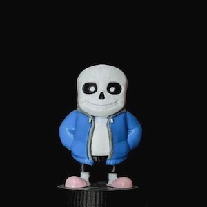 May include: A small figurine of a skeleton character with a white skull, large black eye sockets, and a wide grin. The figure wears a bright blue hooded jacket with grey trim over a white shirt, and has light pink rounded feet on a black base.