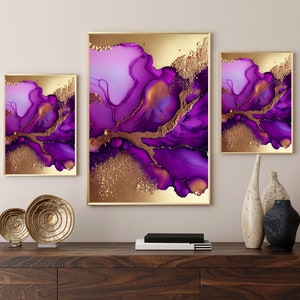 May include: Set of three framed abstract art prints. The artwork features a blend of purple and gold hues, with fluid, ink-like patterns. The gold accents add a touch of luxury. The frames are gold, and the art is displayed on a dark wood surface.