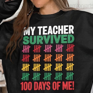 May include: Black sweatshirt featuring the text "MY TEACHER SURVIVED 100 DAYS OF ME!" in white, green, red, orange, and yellow. The design includes tally marks. The word "SURVIVED" is in green, and the phrase "100 DAYS OF ME!" is in red.