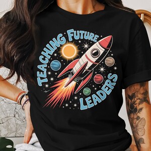 May include: Black t-shirt with a colorful graphic featuring a rocket ship, planets, sun, and stars. The text "TEACHING FUTURE LEADERS" is in a circular design around the rocket. The rocket has red, white, and black details.
