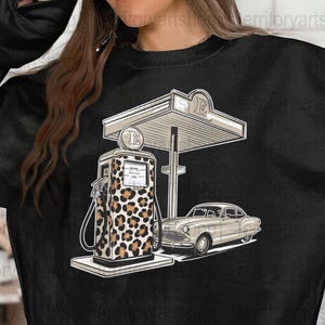 May include: Black sweatshirt with a graphic of a vintage gas station with a leopard print gas pump and a classic car. The gas station sign reads "E".