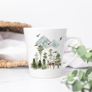 May include: White ceramic mug featuring a watercolor illustration of a forest scene. The design includes green trees, a deer, mountains, and birds. The mug has a curved handle and a tapered shape, suitable for hot beverages.