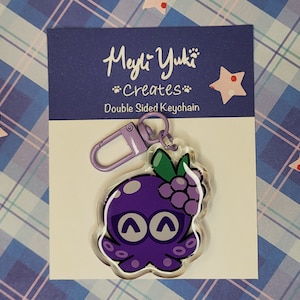 Op de afbeelding: Paarse octopus sleutelhanger met een tros druiven erop. De sleutelhanger is dubbelzijdig en heeft een paarse metalen sluiting. De tekst op de kaart luidt "Meyli-Yuki Creates" en "Double Sided Keychain".