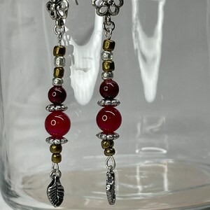May include: A pair of long, silver earrings with red and gold beads. The earrings feature a flower charm at the top and a leaf charm at the bottom.