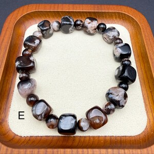 May include: A beaded bracelet featuring a mix of dark brown, black, and cream-colored stones. The bracelet is made of various shapes and sizes of polished beads, strung together on an elastic band. The letter "E" is visible in the image.
