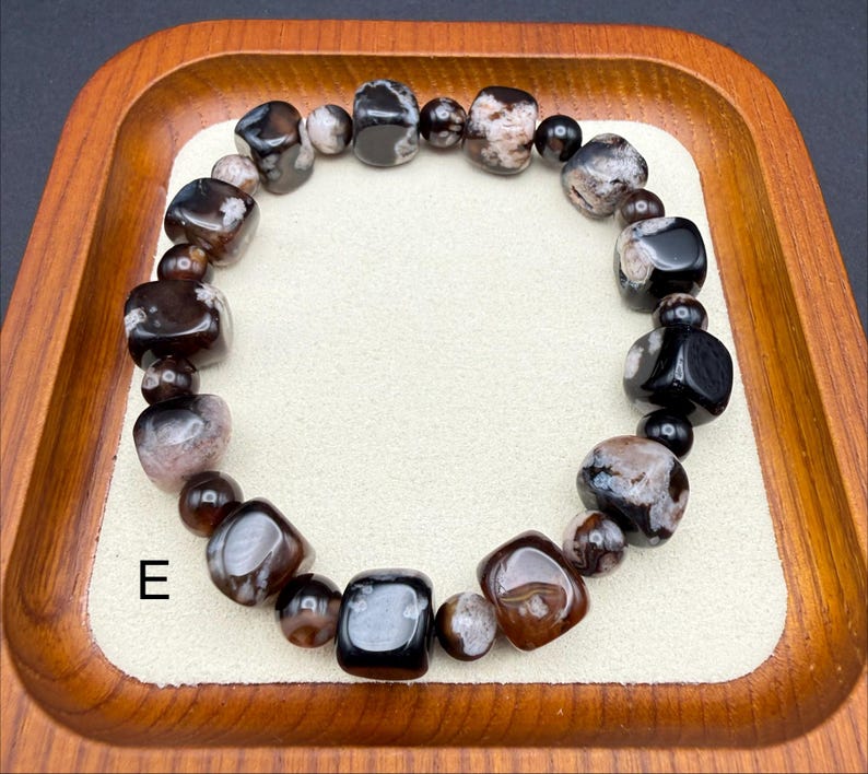 May include: A beaded bracelet featuring a mix of dark brown, black, and cream-colored stones. The bracelet is made of various shapes and sizes of polished beads, strung together on an elastic band. The letter "E" is visible in the image.