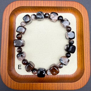 May include: A beaded bracelet featuring a mix of dark brown, black, and gray stones. The bracelet includes both rounded and square-cut beads, creating a unique texture. The letter "E" is visible on the lower left.