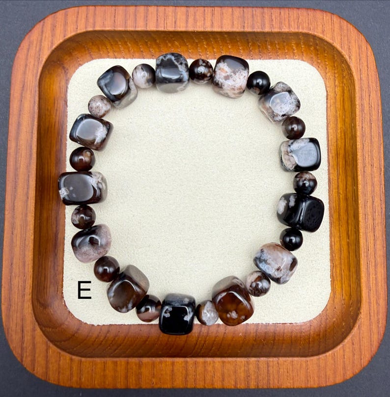 May include: A beaded bracelet featuring a mix of dark brown, black, and gray stones. The bracelet includes both rounded and square-cut beads, creating a unique texture. The letter "E" is visible on the lower left.