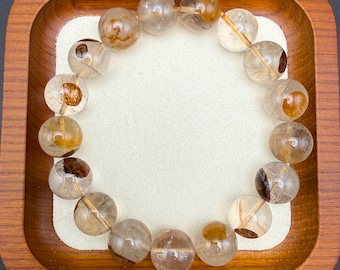 May include: A clear quartz bracelet with inclusions of golden and brown minerals. The round beads are strung together, creating a circular shape. The bracelet is displayed on a white surface inside a wooden tray.