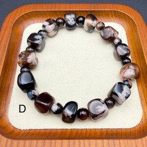 May include: A beaded bracelet featuring a mix of dark brown, black, and white stones. The bracelet is made of various shapes and sizes of polished beads, strung together on an elastic band. The bracelet is displayed on a wooden tray.