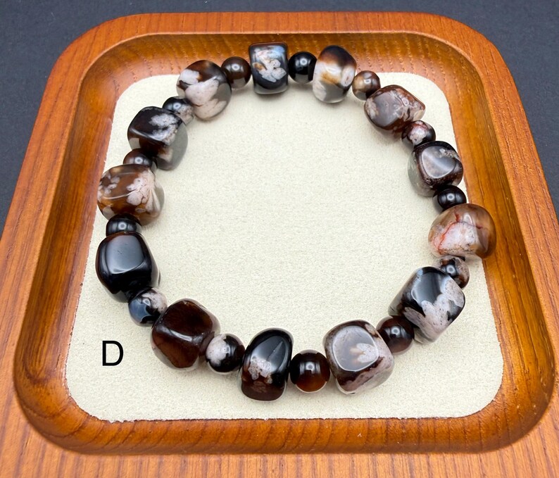 May include: A beaded bracelet featuring a mix of dark brown, black, and white stones. The bracelet is made of various shapes and sizes of polished beads, strung together on an elastic band. The bracelet is displayed on a wooden tray.