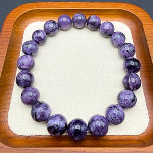 Pu&ograve; includere: Un braccialetto composto da perline rotonde e lucide di charoite viola. Le perline presentano un motivo a vortice di viola scuro e nero. Il braccialetto &egrave; esposto su una superficie bianca all'interno di un vassoio di legno.