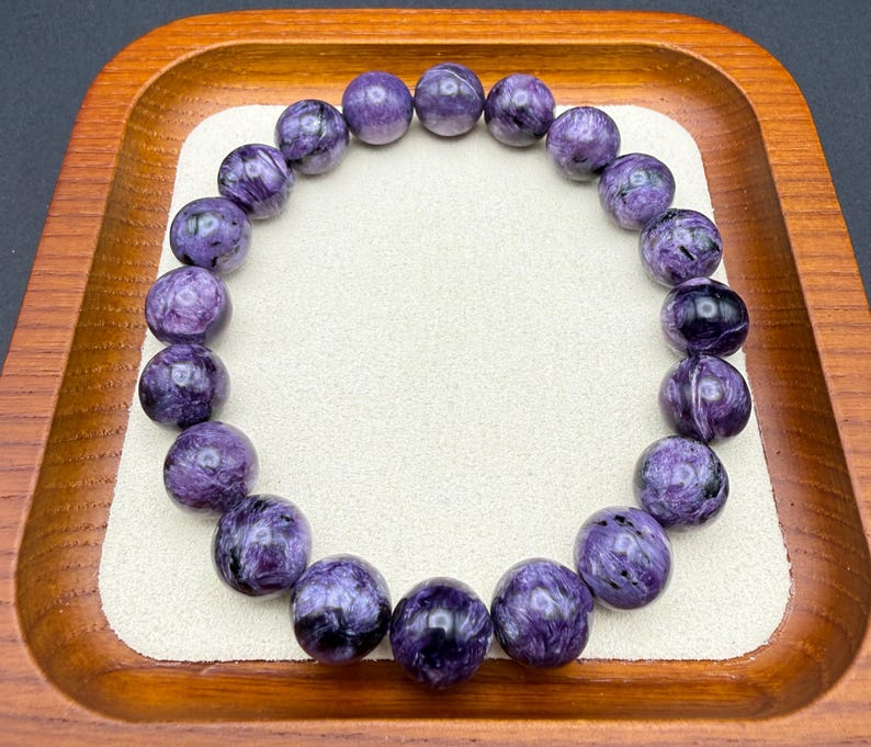 Pu&ograve; includere: Un braccialetto composto da perline rotonde e lucide di charoite viola. Le perline presentano un motivo a vortice di viola scuro e nero. Il braccialetto &egrave; esposto su una superficie bianca all'interno di un vassoio di legno.