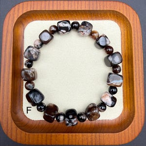 May include: A beaded bracelet featuring a mix of dark brown, black, and cream-colored stones. The bracelet includes both rounded and square-cut beads, creating a unique texture. The letter "F" is visible on the lower left side of the image.