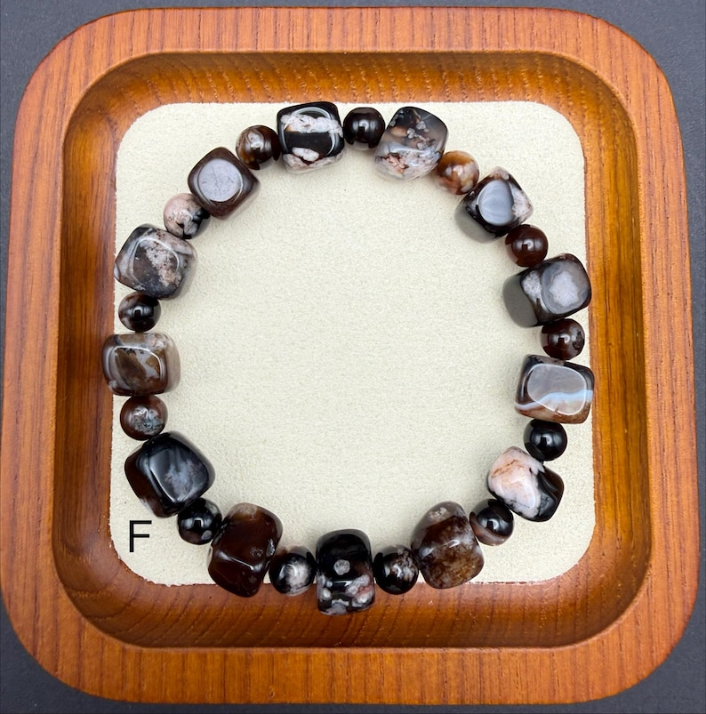 May include: A beaded bracelet featuring a mix of dark brown, black, and cream-colored stones. The bracelet includes both rounded and square-cut beads, creating a unique texture. The letter "F" is visible on the lower left side of the image.