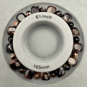 May include: A beaded bracelet with dark brown and black stones, displayed inside a gray measuring tool. The tool indicates a diameter of 6 1/2 inches. The bracelet is made of polished, round beads.
