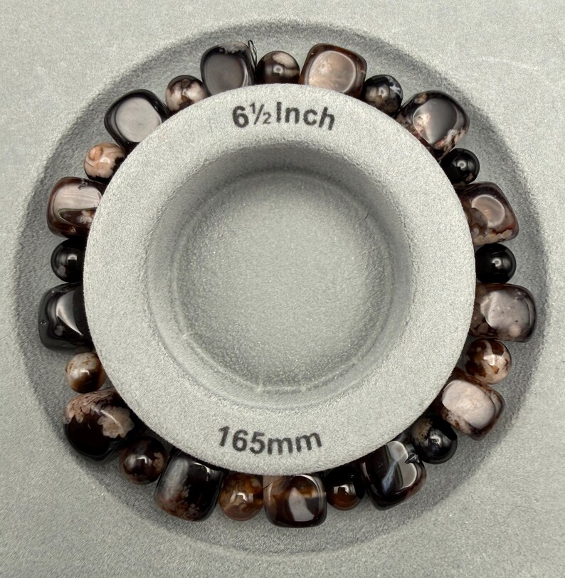 May include: A beaded bracelet with dark brown and black stones, displayed inside a gray measuring tool. The tool indicates a diameter of 6 1/2 inches. The bracelet is made of polished, round beads.