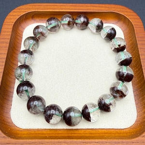 May include: A beaded bracelet featuring clear, round beads with dark brown and green inclusions. The bracelet is displayed on a white surface within a wooden tray. The beads are approximately 0.5 inches in diameter.