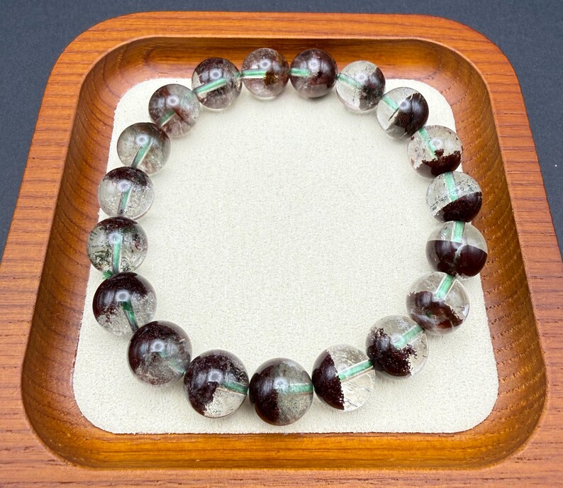 May include: A beaded bracelet featuring clear, round beads with dark brown and green inclusions. The bracelet is displayed on a white surface within a wooden tray. The beads are approximately 0.5 inches in diameter.