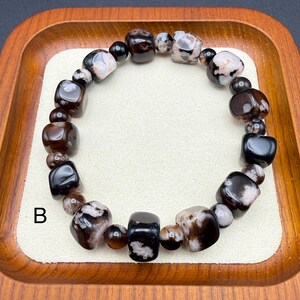May include: A beaded bracelet featuring a mix of dark brown, black, and white patterned stones. The bracelet is composed of various shapes, including rounded and rectangular beads, and is displayed on a cream-colored surface within a wooden tray.