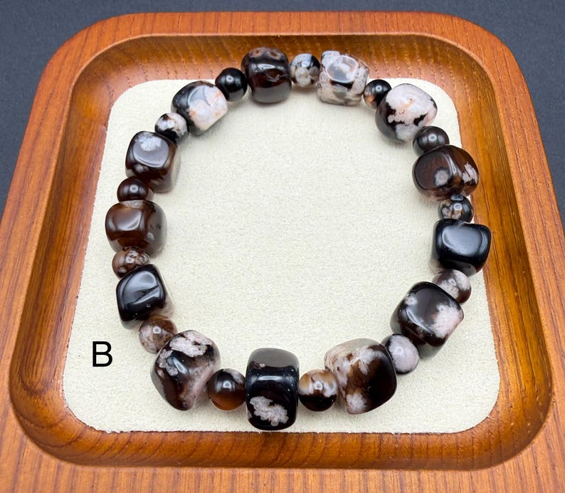 May include: A beaded bracelet featuring a mix of dark brown, black, and white patterned stones. The bracelet is composed of various shapes, including rounded and rectangular beads, and is displayed on a cream-colored surface within a wooden tray.