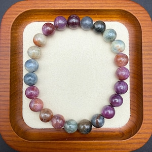 May include: A circular beaded bracelet featuring polished, spherical stones in a gradient of colors including deep purple, dark blue, muted green, and reddish-brown tones, arranged in a continuous loop on a light-colored surface.