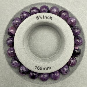Pu&ograve; includere: Un braccialetto di perline rotonde di charoite viola circonda uno strumento di misurazione grigio. Lo strumento &egrave; contrassegnato con "6 1/2 Inch" e "165mm". Le perline hanno un motivo a vortice di viola e bianco.