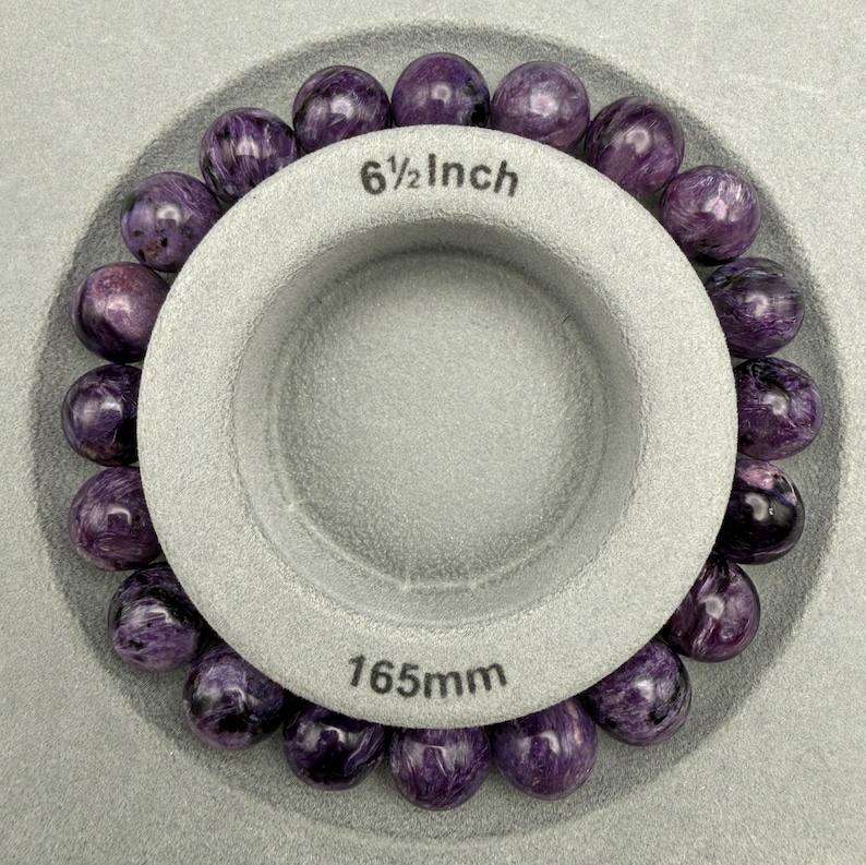 Pu&ograve; includere: Un braccialetto di perline rotonde di charoite viola circonda uno strumento di misurazione grigio. Lo strumento &egrave; contrassegnato con "6 1/2 Inch" e "165mm". Le perline hanno un motivo a vortice di viola e bianco.