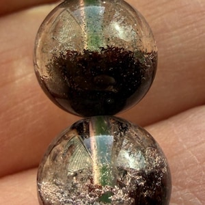 May include: Close-up of a bracelet featuring clear, round beads with dark, textured inclusions. The beads have a reflective, polished surface, and the bracelet is held by a person's hand.