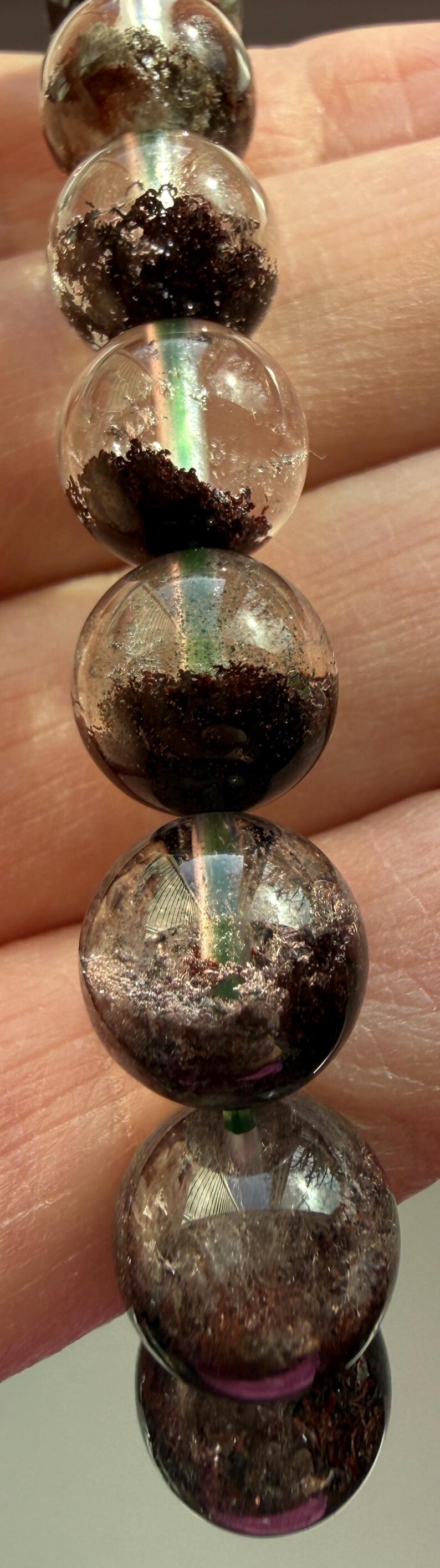 May include: Close-up of a bracelet featuring clear, round beads with dark, textured inclusions. The beads have a reflective, polished surface, and the bracelet is held by a person's hand.