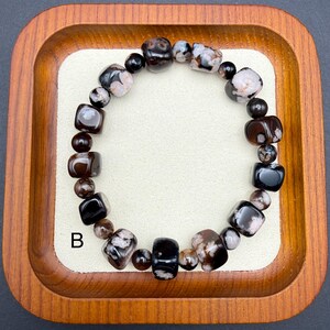 May include: A beaded bracelet featuring a mix of dark brown, black, and speckled white stones. The bracelet is composed of various shapes, including rounded beads and square-cut stones. The letter "B" is visible on the lower left.