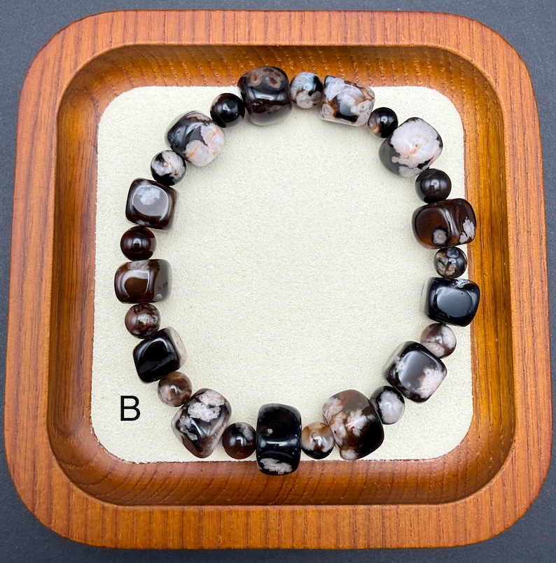 May include: A beaded bracelet featuring a mix of dark brown, black, and speckled white stones. The bracelet is composed of various shapes, including rounded beads and square-cut stones. The letter "B" is visible on the lower left.
