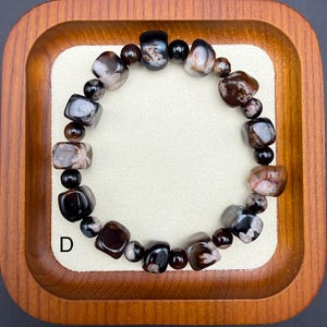 May include: A beaded bracelet featuring a mix of polished, dark brown, black, and gray stones. The bracelet is composed of various shapes, including rounded and rectangular beads. The letter "D" is visible in the lower left corner.