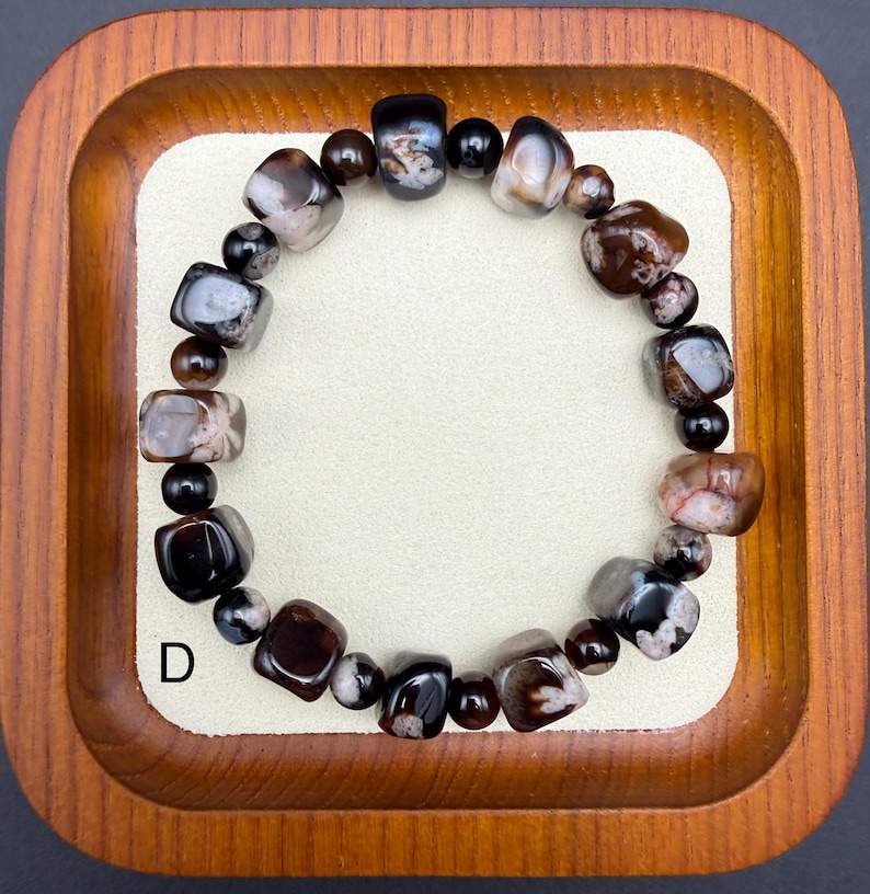 May include: A beaded bracelet featuring a mix of polished, dark brown, black, and gray stones. The bracelet is composed of various shapes, including rounded and rectangular beads. The letter "D" is visible in the lower left corner.