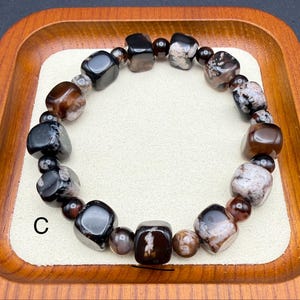 May include: A beaded bracelet featuring a variety of polished stones in shades of brown, black, and white. The bracelet is composed of irregularly shaped beads and smaller, round beads. The letter "C" is visible on the lower left side of the image.