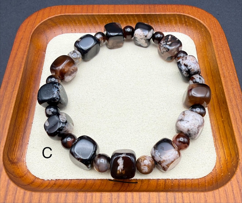 May include: A beaded bracelet featuring a variety of polished stones in shades of brown, black, and white. The bracelet is composed of irregularly shaped beads and smaller, round beads. The letter "C" is visible on the lower left side of the image.
