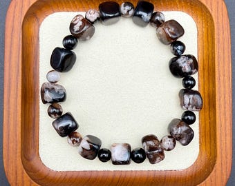 May include: A beaded bracelet featuring a mix of polished, dark brown, black, and speckled stones. The bracelet is composed of various shapes, including rounded and square beads, strung together. The bracelet is displayed on a cream-colored surface within a wooden tray.