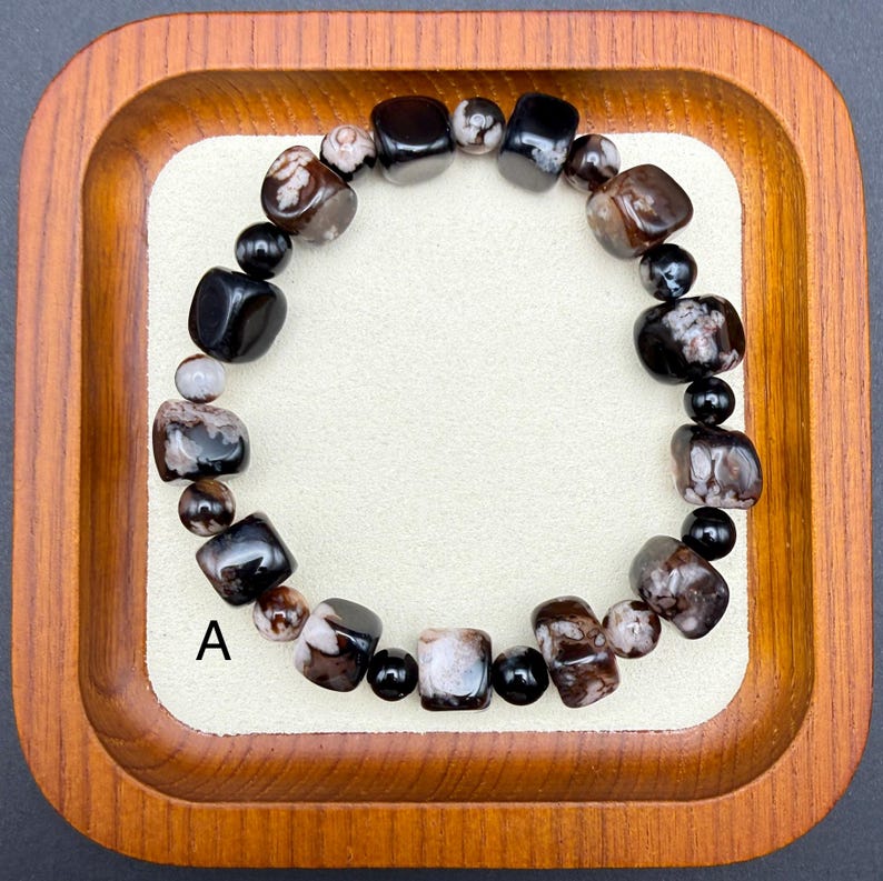 May include: A beaded bracelet featuring a mix of dark brown, black, and cream-colored stones. The bracelet includes both rounded and square-shaped beads, with a mix of polished and matte finishes. The letter "A" is visible on the background.