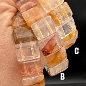 May include: A close-up of a bracelet made of rectangular, translucent, orange-brown gemstones. The bracelet is held against a dark background. The gemstones have internal striations and variations in color.