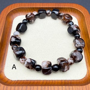 May include: A beaded bracelet featuring a mix of black, brown, and white stones. The bracelet is composed of various shapes and sizes of beads, including rounded and rectangular ones. The bracelet is displayed on a cream-colored surface inside a wooden tray.